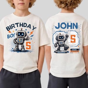 May include: An off-white t-shirt featuring a graphic of a robot, the text BIRTHDAY BOY, and the number 5 on the front. The back displays the name JOHN, a robot graphic, the number 5, and the text MODE ACTIVATED in a blue and orange color scheme.