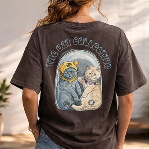 May include: Dark gray t-shirt featuring a graphic of two cats inside a clear capsule. One cat wears a yellow tiger hat, the other a beaded collar. The text "The Cat Collective" arches above the cats.