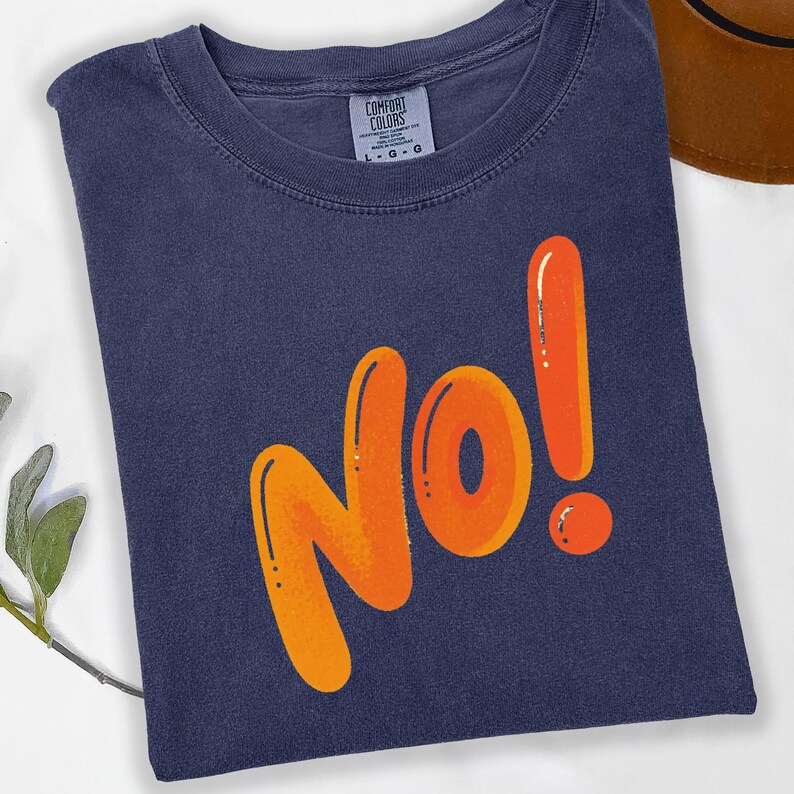 May include: A navy blue t-shirt with the word "No!" in large orange bubble letters. The shirt has a crew neck and the Comfort Colors brand tag is visible. The shirt is folded on a white surface.