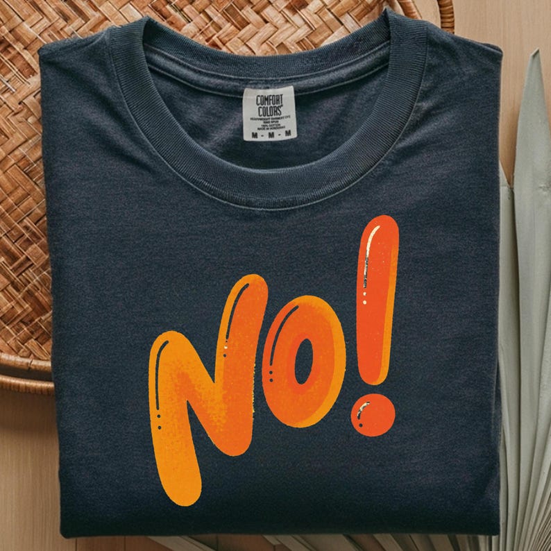 May include: Dark blue t-shirt with the word "No!" in large, orange, bubble letters. The shirt has a crew neck and a tag that reads "Comfort Colors". The shirt is folded neatly on a woven surface.