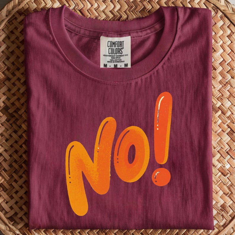 May include: A maroon t-shirt with the word "No!" in large, orange, bubble letters. The shirt has a crew neck and a tag that reads "Comfort Colors". The shirt is folded on a woven surface.