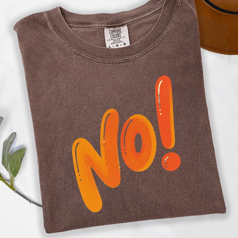 May include: A brown t-shirt with the word "No!" in orange bubble letters. The shirt has a crew neck and a tag that reads "Comfort Colors". A brown hat is partially visible in the background.
