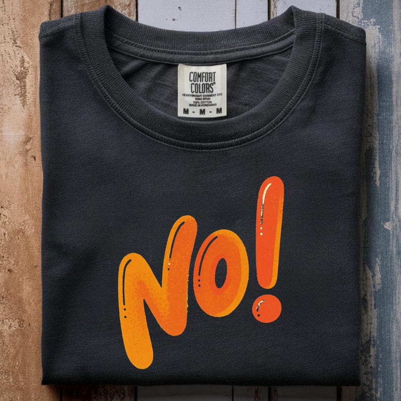 May include: A dark gray t-shirt with the word "No!" in orange, stylized lettering. The exclamation point is a bright orange. The shirt has a crew neck and a tag that reads "Comfort Colors".