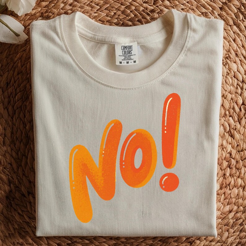 May include: Cream-colored t-shirt with the word "No!" in orange bubble letters. The shirt has a crew neck and a tag that reads "Comfort Colors". The t-shirt is folded on a woven surface.