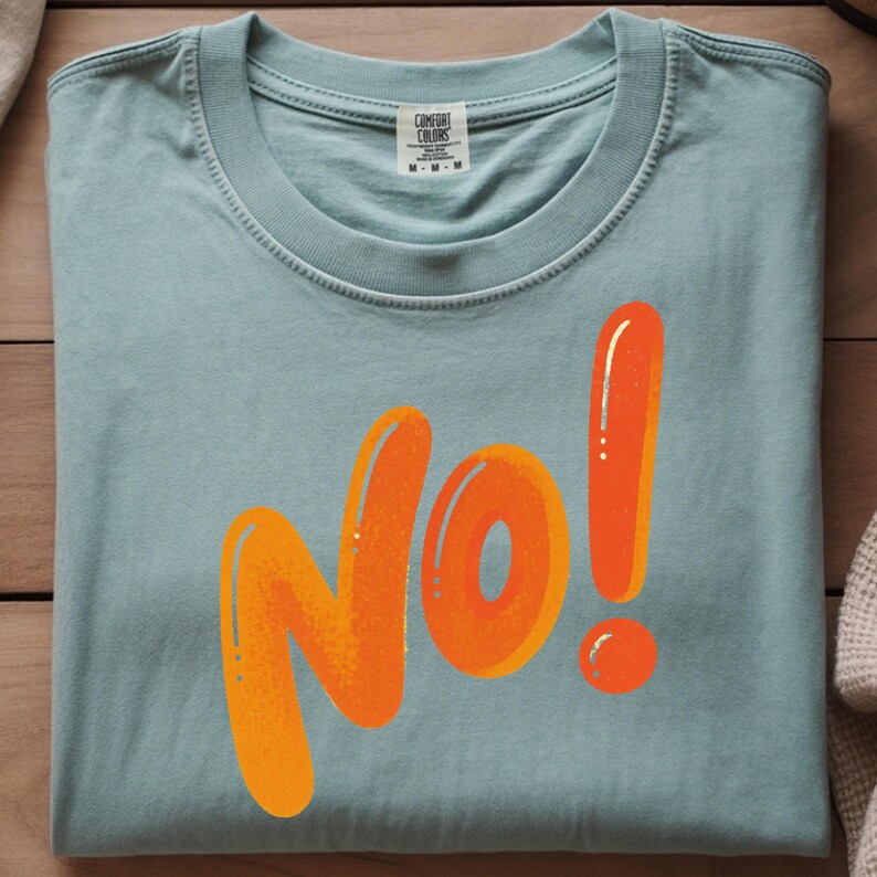 May include: A light blue t-shirt with the word "NO!" in large orange bubble letters. The shirt is folded and has a tag at the neckline. The graphic has a gradient effect.