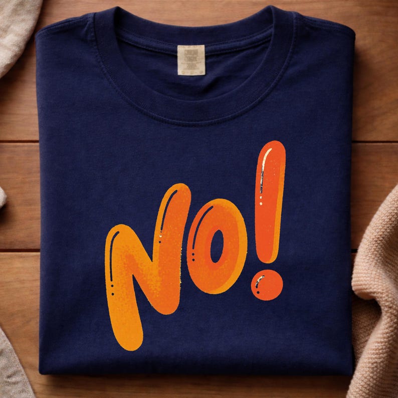 May include: Navy blue t-shirt with the word "No!" in orange, outlined in a darker orange. The exclamation point is a vertical line with a small orange circle at the bottom. The shirt is folded neatly on a wooden surface.