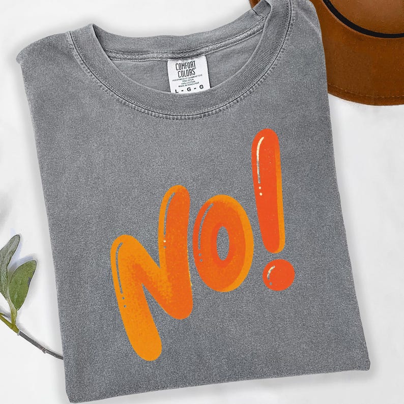 May include: A gray Comfort Colors t-shirt with the word "NO!" in large orange bubble letters. The shirt is folded and has a tag at the neckline. A brown hat is partially visible in the background.