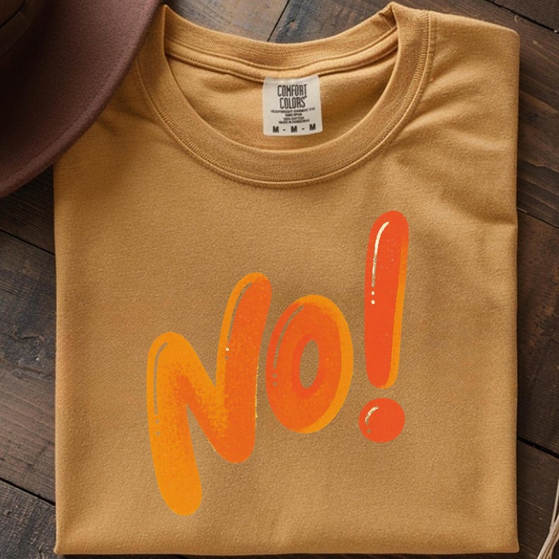 May include: Mustard-colored t-shirt with the word "No!" in orange, outlined in a darker orange. The shirt has a crew neck and the brand "Comfort Colors" is visible on the tag.