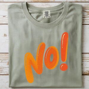 May include: Sage green t-shirt with the word "NO!" in orange bubble letters. The t-shirt has a crew neck and short sleeves. The tag reads "Comfort Colors".