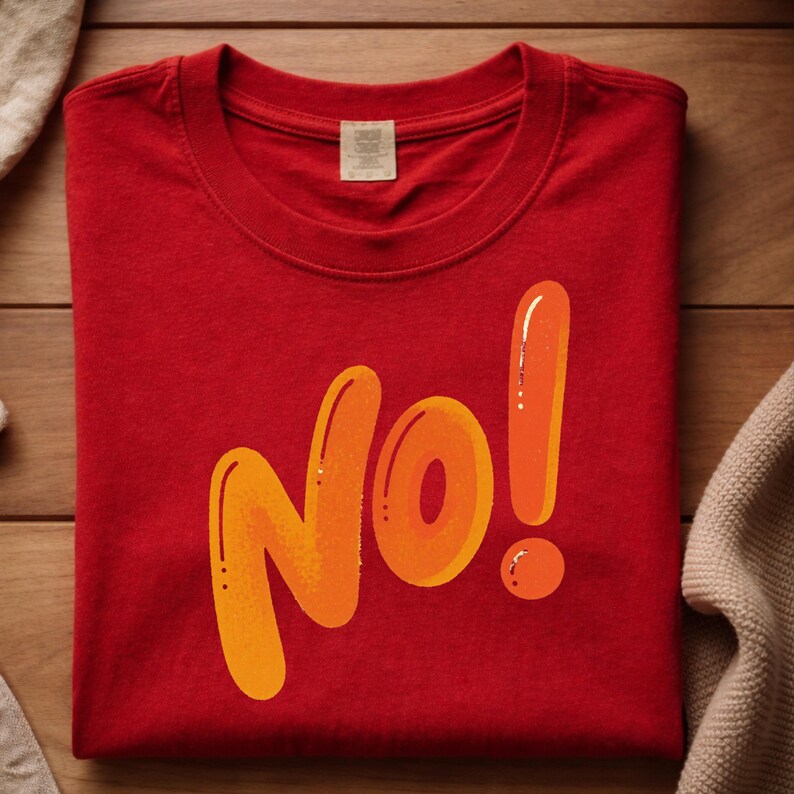 May include: A red t-shirt with the word "No!" in orange and yellow bubble letters. The t-shirt is folded neatly on a wooden surface, with a neutral-colored fabric on either side.