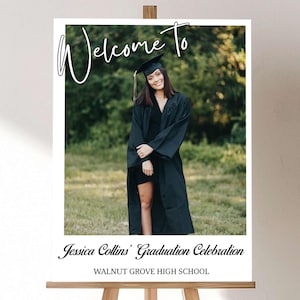 May include: A graduation welcome sign on a wooden easel. The sign features a photo of a graduate in a cap and gown, with the words "Welcome To" in script. Below the photo, it reads "Jessica Collins' Graduation Celebration" and "Walnut Grove High School."