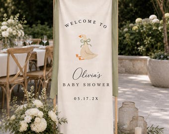 May include: A vertical off-white fabric banner suspended on a wooden frame with sage green ribbons. The banner features a graphic of a goose with a green bow, and black text reading "WELCOME TO", "Olivia's", "BABY SHOWER", and "05.17.2X".