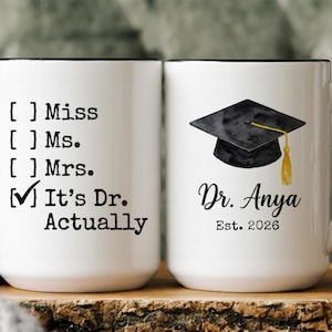 May include: White ceramic mug with black handle and rim. One side reads "It's Dr. Actually" with checkmark. The other side features a graduation cap and "Dr. Anya Est. 2026". A perfect gift for a new doctor.