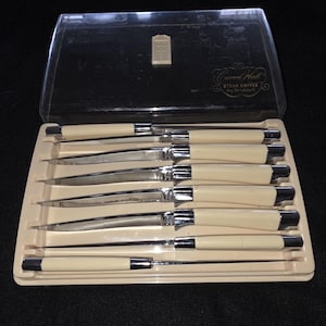 Può includere: Un set di coltelli da bistecca in una custodia di plastica trasparente. I coltelli hanno manici color crema con ghiere e lame argentate. La custodia è etichettata "Carvel Hall Steak Knives by Bridell".