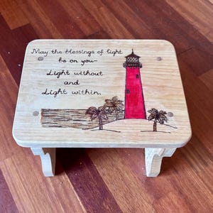 Op de afbeelding: Een houten opstapje met een handgeschilderde vuurtorenscène. De rode vuurtoren staat centraal, met palmbomen en oceaandetails. De kruk heeft de tekst "May the blessings of light be on you - Light without and Light within."