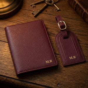Può includere: Set di portapassaporto e targhetta per bagagli in pelle bordeaux con finiture color oro. Entrambi gli articoli sono impressi con le iniziali "M.R." nell'angolo in basso a destra. Il set è esposto su una superficie di legno.