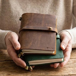 May include: Two leather journals, one brown and one green, held by a person. The brown journal has a strap closure and the initials "MK". The green journal has the initial "T". The journals are on a wooden surface.