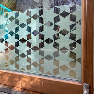 May include: A window with a decorative film featuring a repeating pattern of stars and diamond shapes in shades of brown, gray, and white. The window is framed by a wooden border.