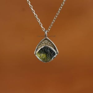 May include: A silver pendant necklace featuring a triangular pendant with a green and black stone. The pendant is suspended from a delicate silver chain. The pendant has a decorative design with crescent moon shapes.