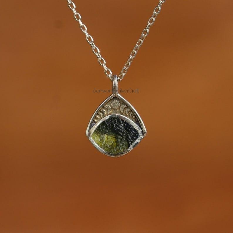 May include: A silver pendant necklace featuring a triangular pendant with a green and black stone. The pendant is suspended from a delicate silver chain. The pendant has a decorative design with crescent moon shapes.