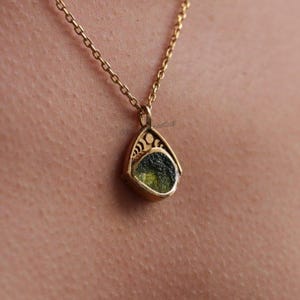 May include: A gold-colored pendant necklace with a teardrop-shaped pendant. The pendant features a green stone set within a decorative gold frame. The necklace chain is also gold. The pendant is approximately 0.75 inches in length.