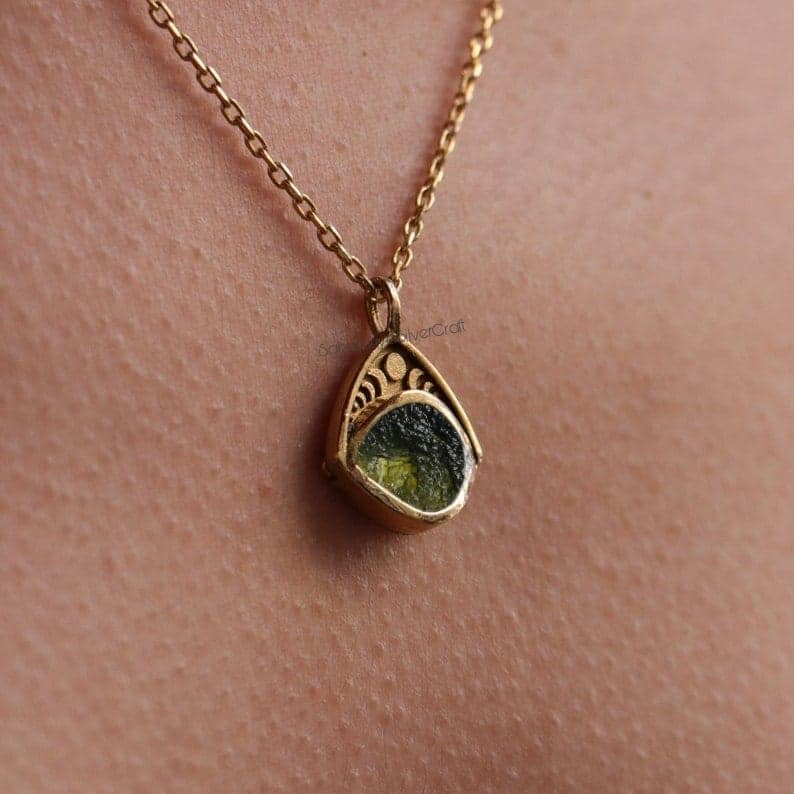 May include: A gold-colored pendant necklace with a teardrop-shaped pendant. The pendant features a green stone set within a decorative gold frame. The necklace chain is also gold. The pendant is approximately 0.75 inches in length.