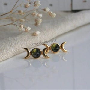 Op de afbeelding: Een paar goudkleurige oorstekers met een halve maan ontwerp en een cirkelvormige steen met groen-zwarte vlekken. De oorbellen worden gepresenteerd op een wit oppervlak. Sieraden.