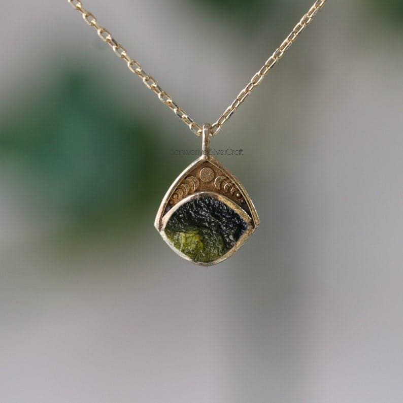 May include: A gold-colored pendant necklace featuring a unique, irregularly shaped green and black stone. The pendant has a decorative, geometric design with small circles. The necklace chain is also gold-colored.