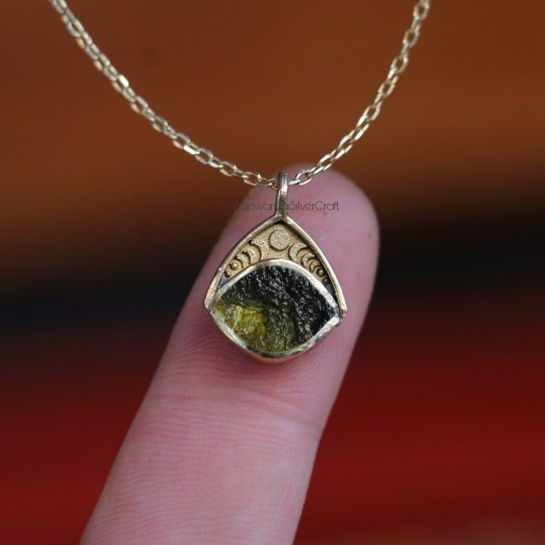 May include: A gold-colored pendant necklace featuring a triangular pendant with a green and black stone. The pendant is adorned with crescent moon and swirl designs. The necklace is displayed against a blurred background.