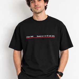 Op de afbeelding: Een zwart T-shirt met korte mouwen en een horizontale rode lijn over de borst. Boven de lijn staat witte tekst: 5-hour limit Resets in 4 hr 59 min 100%. Het T-shirt heeft een ronde hals en een relaxte pasvorm van effen zwarte stof.