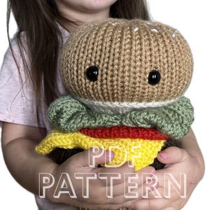 May include: A handmade, plush burger toy with a brown bun, green lettuce, red tomato, and yellow cheese. The burger has black button eyes and is held by a person. The text "PDF PATTERN" is overlaid on the image.