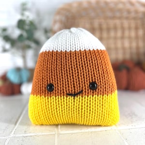 Puede incluir: Un peluche de maíz de caramelo tejido con una cara sonriente. El juguete presenta secciones blancas, naranjas y amarillas, con ojos de botón negros y una boca cosida. El peluche es una decoración con temática de Halloween.