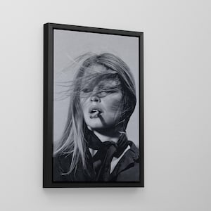 May include: Monochrome framed print featuring a woman with long blonde hair and a cigarette. She wears a dark top and scarf. The portrait captures a moment with wind-blown hair.