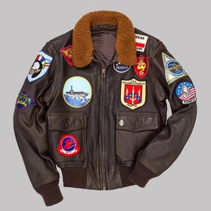 Puede incluir: Chaqueta bomber de cuero marrón oscuro con cuello de borrego marrón. La chaqueta presenta múltiples parches con temas militares y náuticos, incluyendo los textos "SEABEES" y "TOMCAT". Tiene dos bolsillos delanteros y dobladillo y puños acanalados.