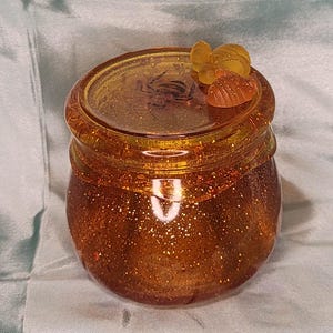 May include: A small, round, orange-tinted jar with a glittery finish. The lid features a small, amber-colored insect and decorative embellishments. The jar is likely for storing small items or as a decorative piece.