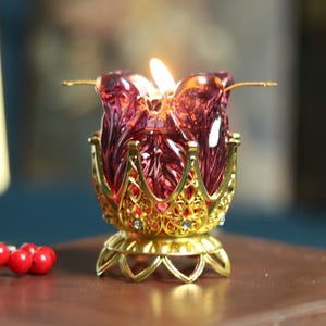 Può includere: Un portacandele in vetro rosso rubino a forma di fiore di loto acceso, posto su una base in filigrana dorata. Il portacandele è appoggiato su un libro rilegato in pelle marrone, con una fila di perline rosse nelle vicinanze.