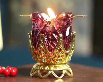 May include: A lit, ruby-colored glass candle holder in the shape of a lotus flower, set in a gold-colored filigree base. The candle holder sits on a brown leather-bound book, with a string of red beads nearby.