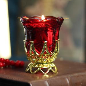 以下が含まれることがあります： 金色のフィリグリーの台座とアクセントが付いた、火の灯った赤いガラスのキャンドルホルダー。茶色の革張りの本の上に置かれています。赤いビーズの列が前景に見え、宗教的なテーマを示唆しています。