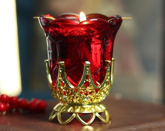 May include: A lit red glass candle holder with a gold filigree base and accents, resting on a brown leather-bound book. A string of red beads is visible in the foreground, suggesting a religious theme.