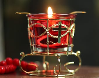 May include: A lit red candle in a gold-colored metal holder with a cross design. The candle sits on a brown leather-bound book, with red beads and a blue background.