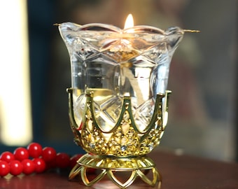 May include: A lit glass candle holder with a gold base sits atop a brown leather-bound book. A red rosary and a framed image are in the background. The candle holder has a clear, faceted glass cup and a gold crown-shaped base.