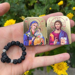 May include: A black knotted prayer rope and two small, gold-colored religious icons are displayed. The icons depict religious figures in vibrant colors. The background features yellow flowers and green foliage.