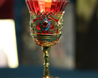May include: A lit candle in a decorative red and gold goblet. The goblet has ornate gold filigree and a jewel-toned centerpiece. The candle flame glows brightly, casting a warm light. The goblet sits on a brown leather-bound book.