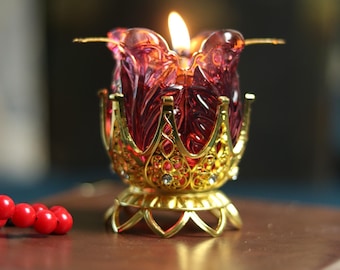 May include: A lit candle in a decorative, ruby-colored glass holder with a gold-colored filigree base. The candle holder has a floral design and is embellished with small, colorful jewels. A string of red beads is in the foreground.