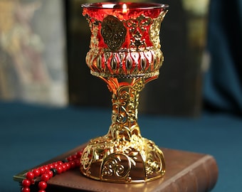 May include: A gold-colored chalice with a red interior, ornate detailing, and a candle inside. The chalice sits atop a closed brown book, with a red rosary and cross in the foreground. The background includes religious artwork.