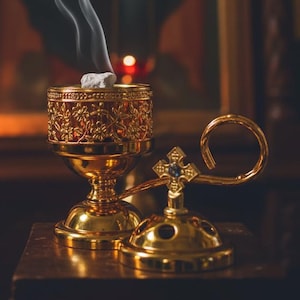 May include: A gold censer with detailed floral patterns, releasing trails of white smoke. The censer sits on a wooden surface, with a blurred religious icon in the background. A small, gold incense holder is also visible.