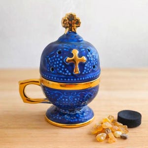 Peut inclure: Un brûleur d'encens en céramique bleu avec des détails dorés et une croix. Le brûleur a une poignée et un couvercle avec un dessus en forme de croix. De l'encens et un disque de charbon sont à côté.