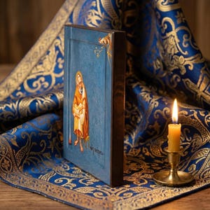 May include: A religious icon featuring a figure in a gold robe against a blue background. The icon is framed in dark wood and rests on a blue and gold patterned fabric. A lit candle in a brass holder is in the foreground.