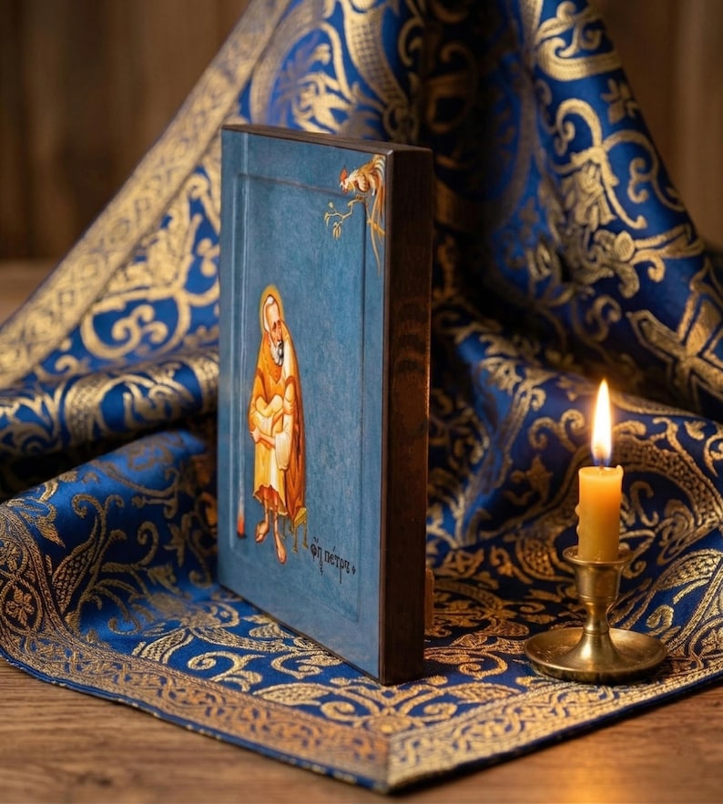 May include: A religious icon featuring a figure in a gold robe against a blue background. The icon is framed in dark wood and rests on a blue and gold patterned fabric. A lit candle in a brass holder is in the foreground.