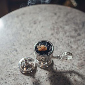 May include: A detailed silver incense burner with a lid and handle, containing dark charcoal and amber resin. Smoke gently rises from the burner, indicating it is lit. The burner is placed on a speckled, light-coloured surface.